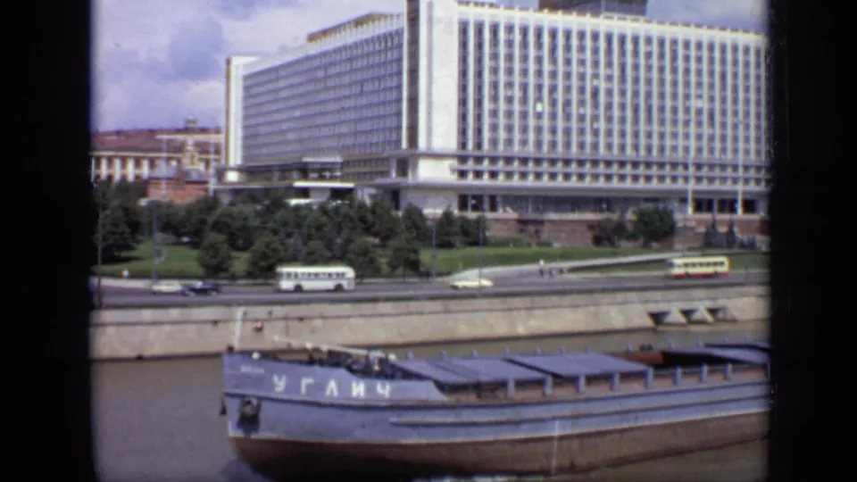 1971 Moscow home-movie frame showing the barge continuing along the river with the embankment, buses, cars, trees, and modernist building behind it.