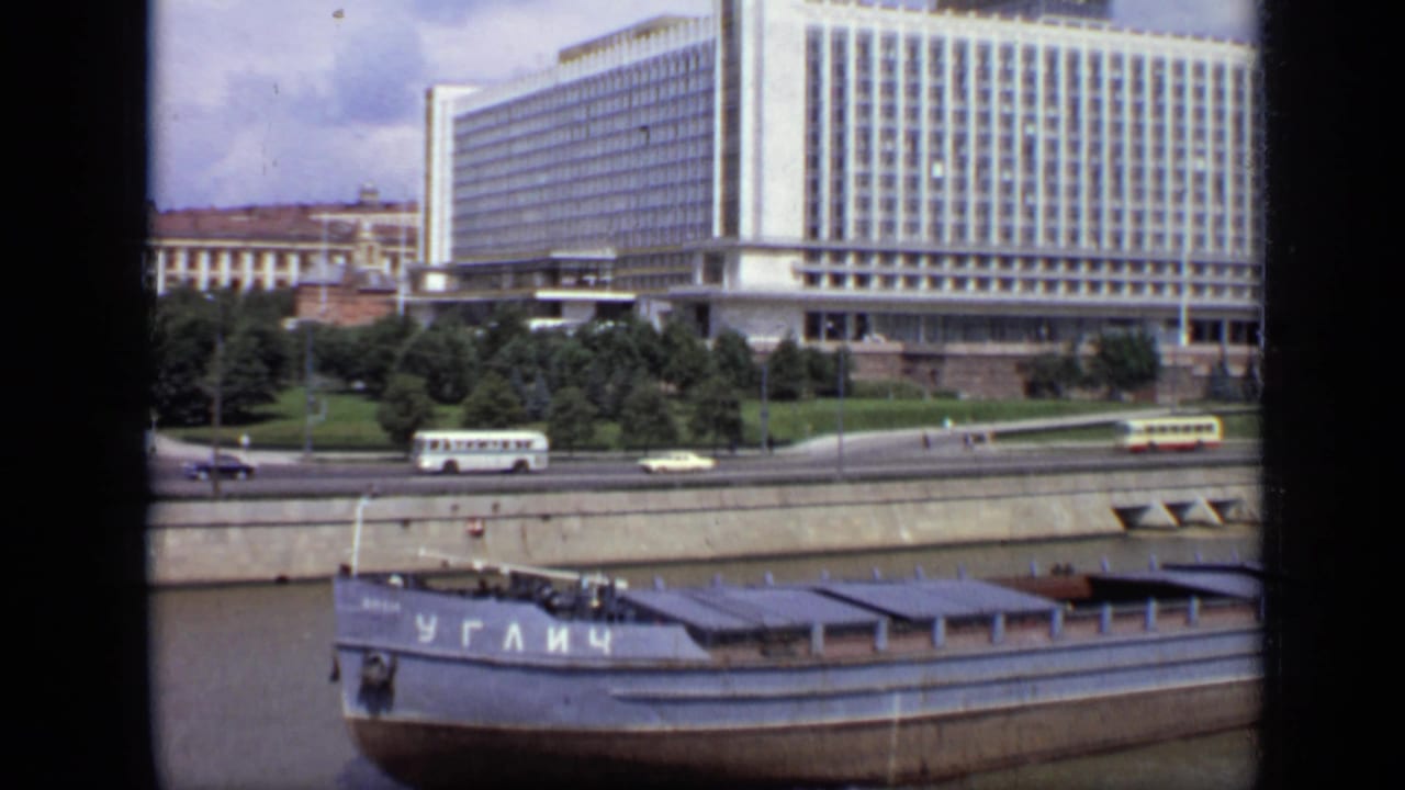 1971 Moscow river view with a white Soviet-era boat, Cyrillic hull lettering, road traffic, trees, and a large riverside building.