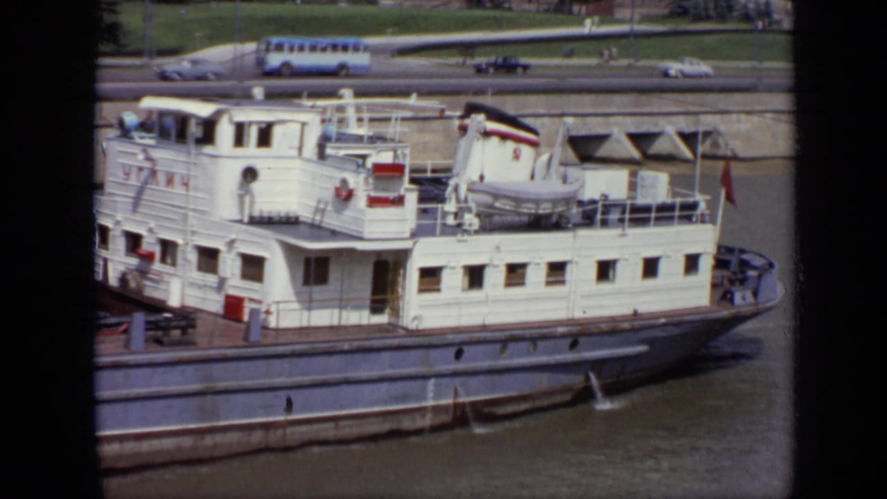 White passenger riverboat in Moscow with cabin windows, deck railings, lifeboat, and distant embankment traffic in 1971 footage.