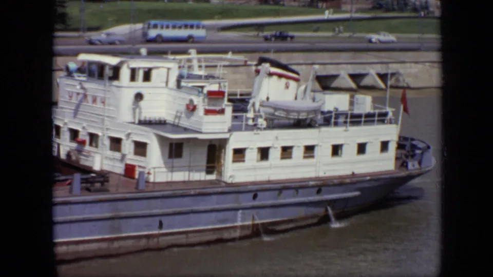 White passenger riverboat in Moscow with cabin windows, deck railings, lifeboat, and distant embankment traffic in 1971 footage.