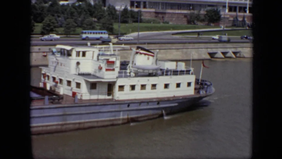 1971 Moscow home-movie frame showing the white riverboat below a stone embankment, with greenery and period road traffic behind.