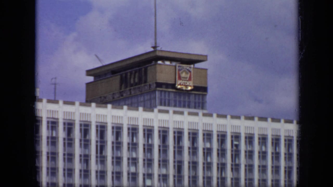 Red-and-white bus parked below a white building with a rooftop emblem.