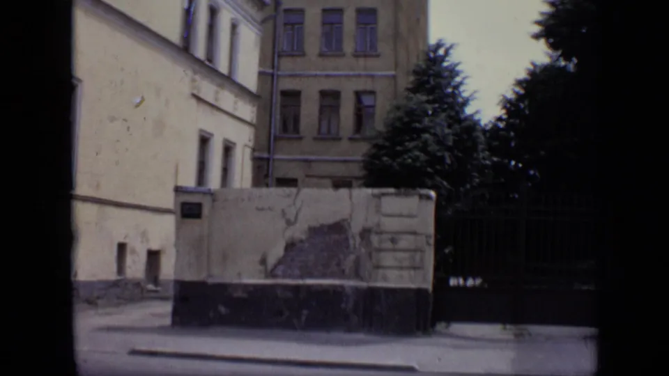 Brief 1971 Moscow street cutaway showing a pale building facade, wall, dark gate, pavement, and trees after the bus view.