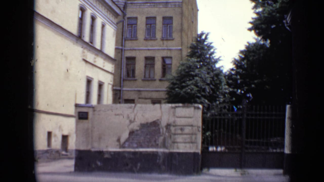 1971 Moscow exterior view showing pale building walls, a weathered wall section, black gate, windows, and leafy trees.