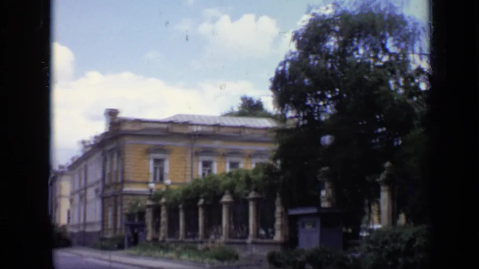 1971 Moscow street exterior with an older low building, columned fence, dark trees, and bright clouded sky.