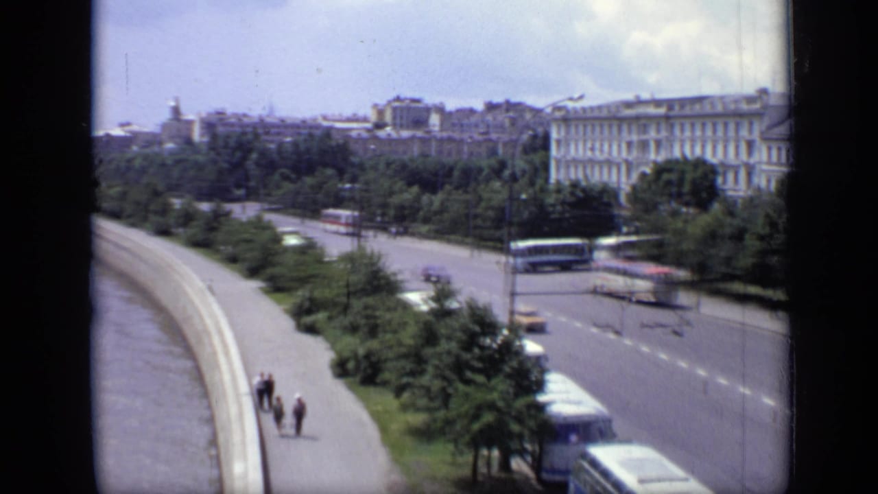 Elevated view of a river embankment with boulevard traffic, pedestrians, trees, and large pale buildings.