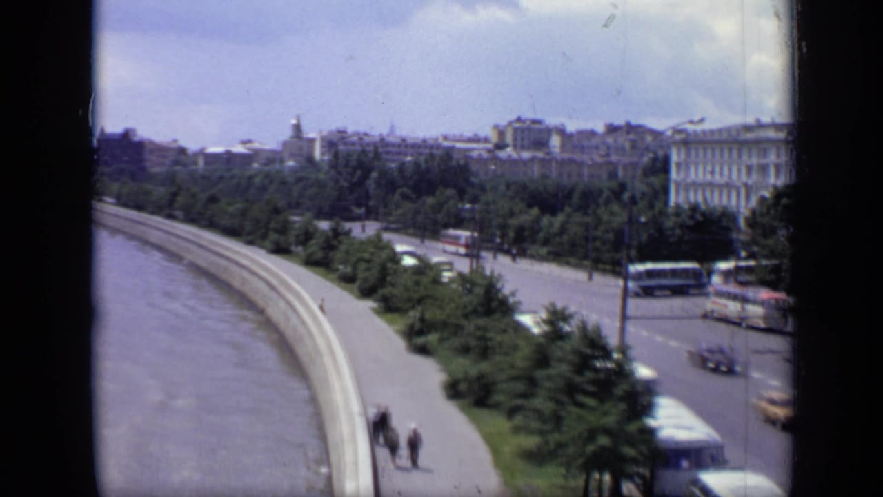 River curve and roadside trees with buses, cars, pedestrians, and city buildings in the distance.
