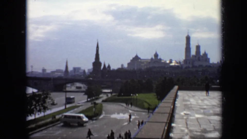 1971 8mm frame of Moscow's Kremlin skyline across the riverfront, with small pedestrians and a vehicle below an overcast sky.