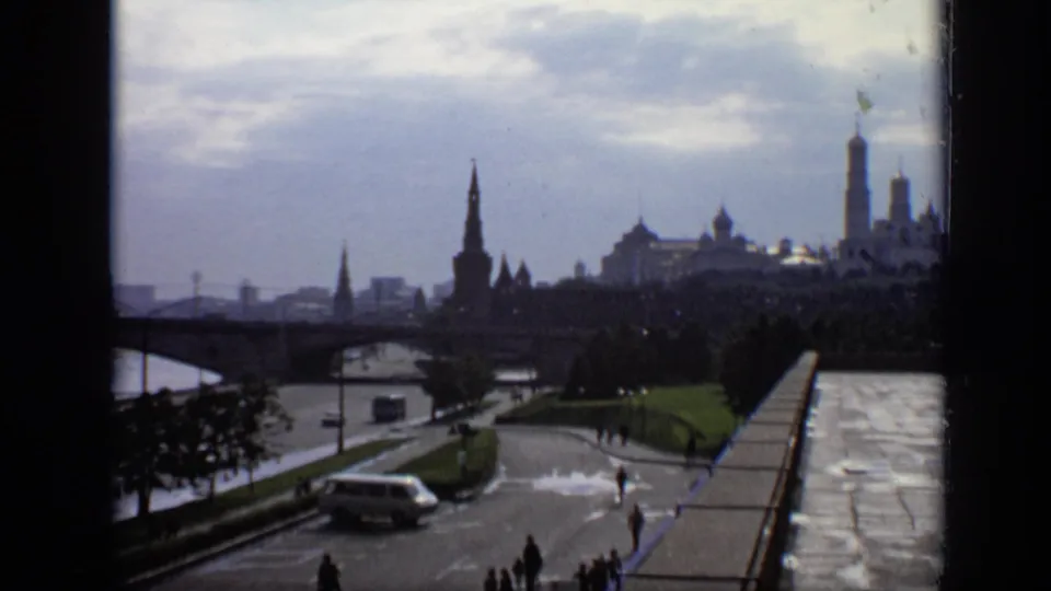 1971 Moscow home-movie view holds on the Kremlin skyline, with foreground pedestrians, a light-colored van, trees, and reflective wet paving.
