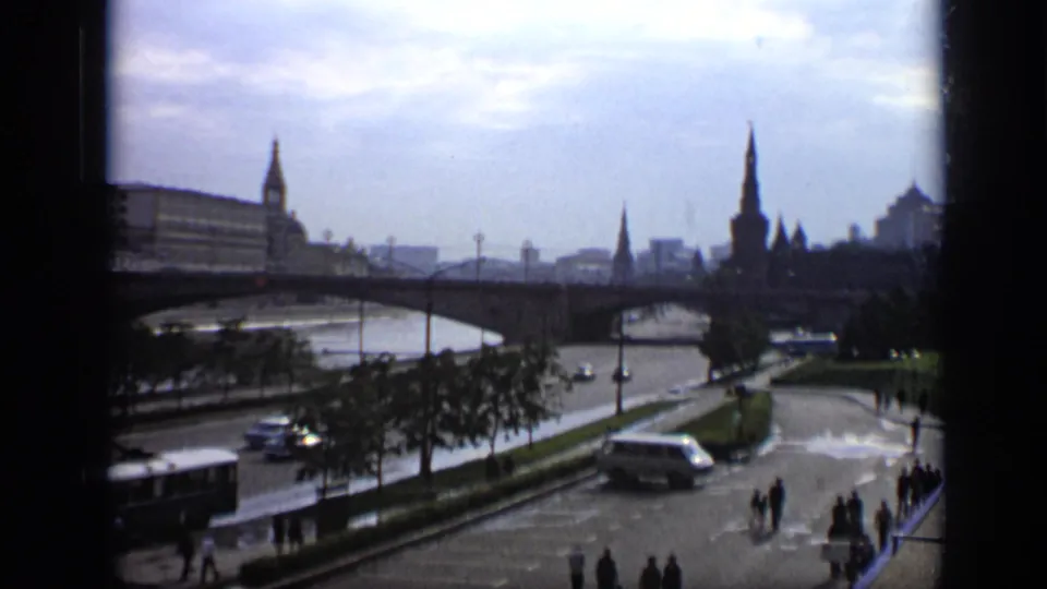 1971 Moscow home-movie view with a river bridge, distant spires, a large vehicle near the embankment, cars, trees, and pedestrians.