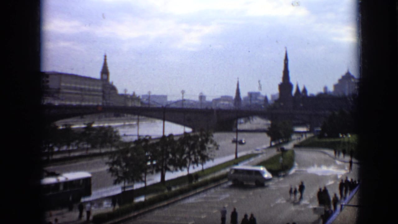 Wide riverfront view with bridge, roadway, pedestrians, and distant city spires under cloudy sky.