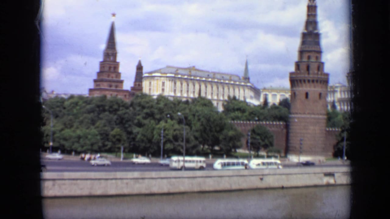 Kremlin towers, pale government building, trees, river embankment, buses, and cars.