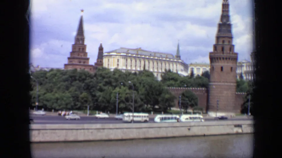 Moscow Kremlin riverfront view with red brick towers, a large pale building, tree line, roadway traffic, and white buses along the embankment.