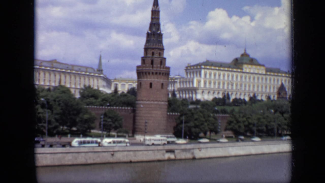 Kremlin tower, pale palace buildings, trees, road traffic, river embankment, and cloudy sky.