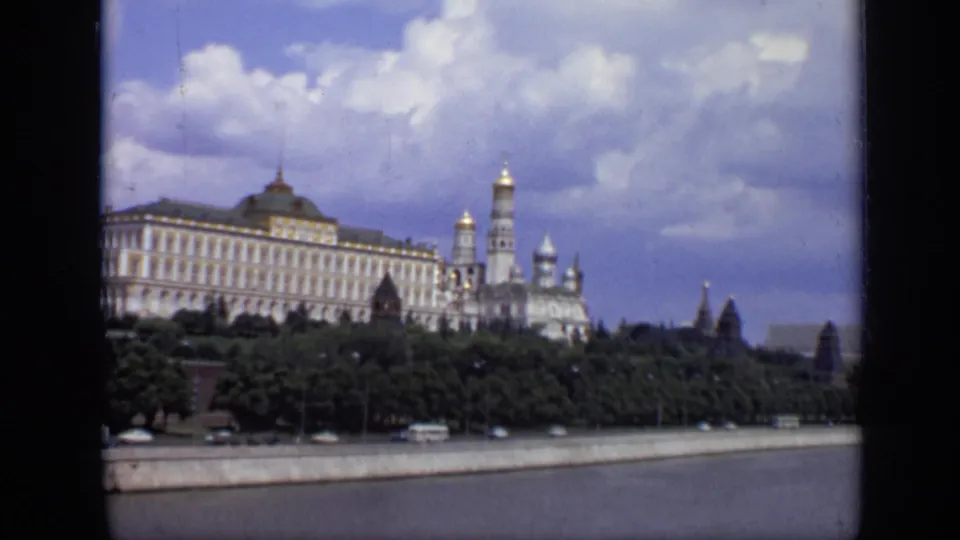 1971 Moscow home-movie frame showing the Kremlin skyline above a green embankment, with river water and passing vehicles below.