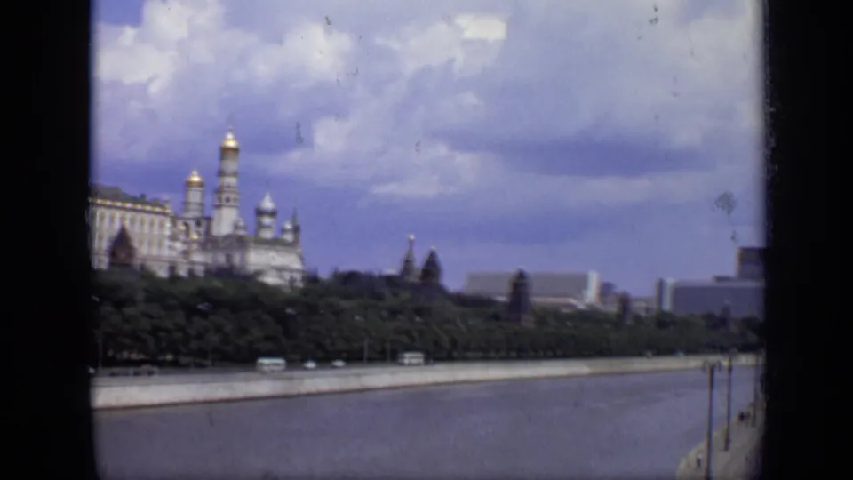 1971 Moscow frame showing Kremlin towers over the riverside trees, with water, embankment traffic, and modern structures in the distance.