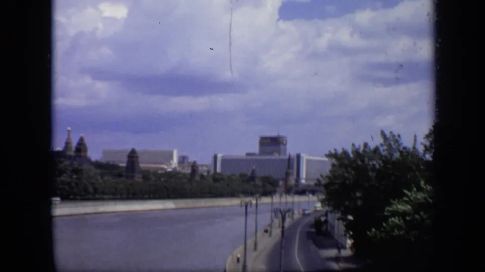 1971 Moscow home-movie frame showing the Moskva River, tree-lined shore, distant towers, modern buildings, and a road following the embankment.