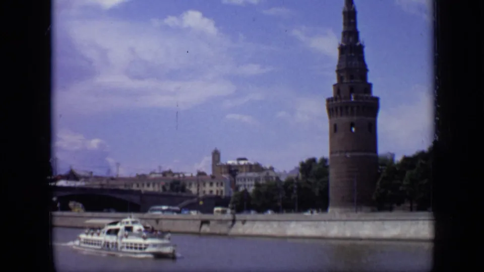 1971 Moscow river view with a prominent Kremlin tower near the embankment, a tour boat on the water, bridge, trees, and riverside buildings.