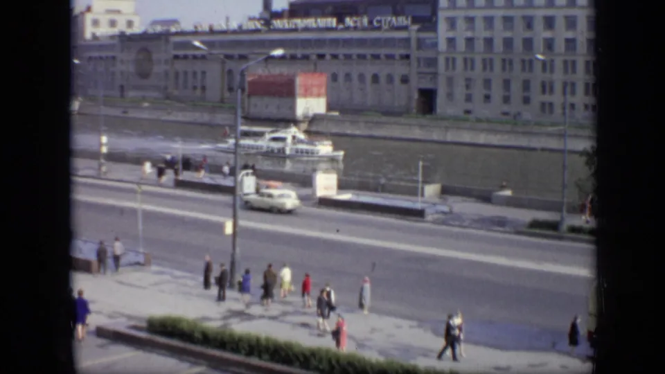 1971 Moscow embankment view showing pedestrians, a white car, a river boat, and a large building with Cyrillic rooftop signage.