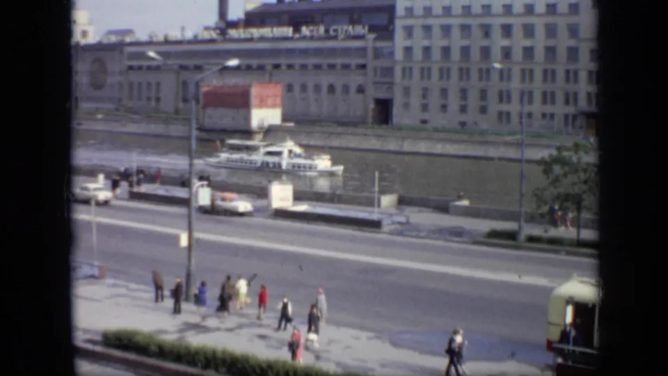 Wide 1971 8mm Moscow riverfront shot with pedestrians near the road, a white car, a boat, and Cyrillic signage across the river.
