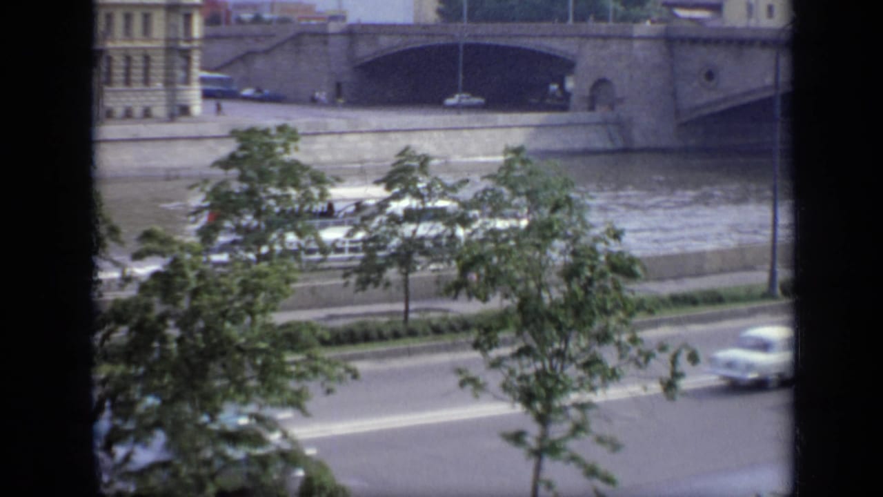 1971 Moscow embankment view with cars passing beneath trees and a large stone bridge spanning the river behind them.