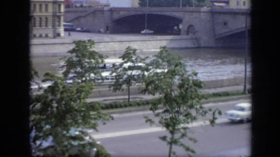 1971 Moscow embankment view with cars passing beneath trees and a large stone bridge spanning the river behind them.