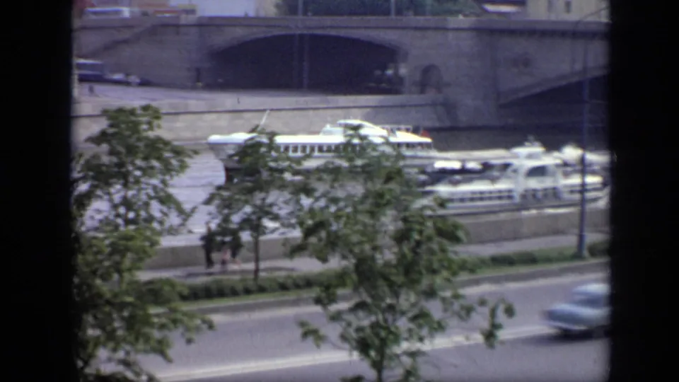 1971 Moscow riverfront with passenger boats partly screened by trees, a stone bridge behind, and cars moving along the road below.