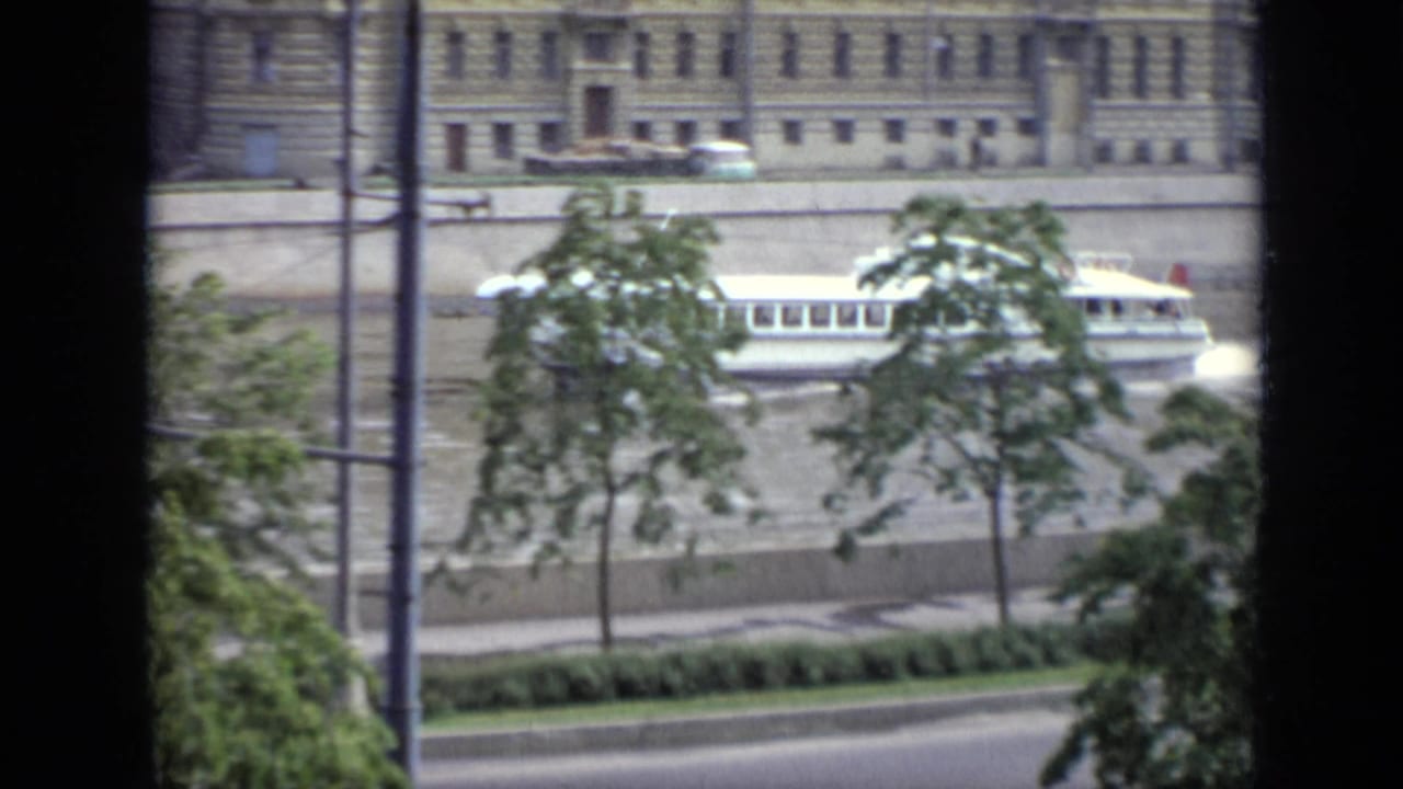 Riverboat continuing past trees and a large riverside building.