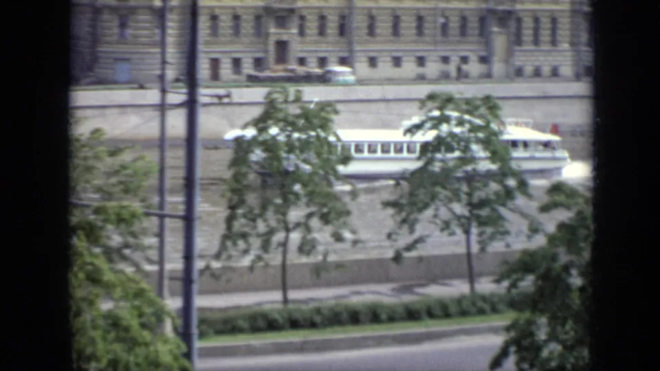 White riverboat cruises rightward on the Moscow waterway, with trees, lampposts, and a formal riverside facade in view.