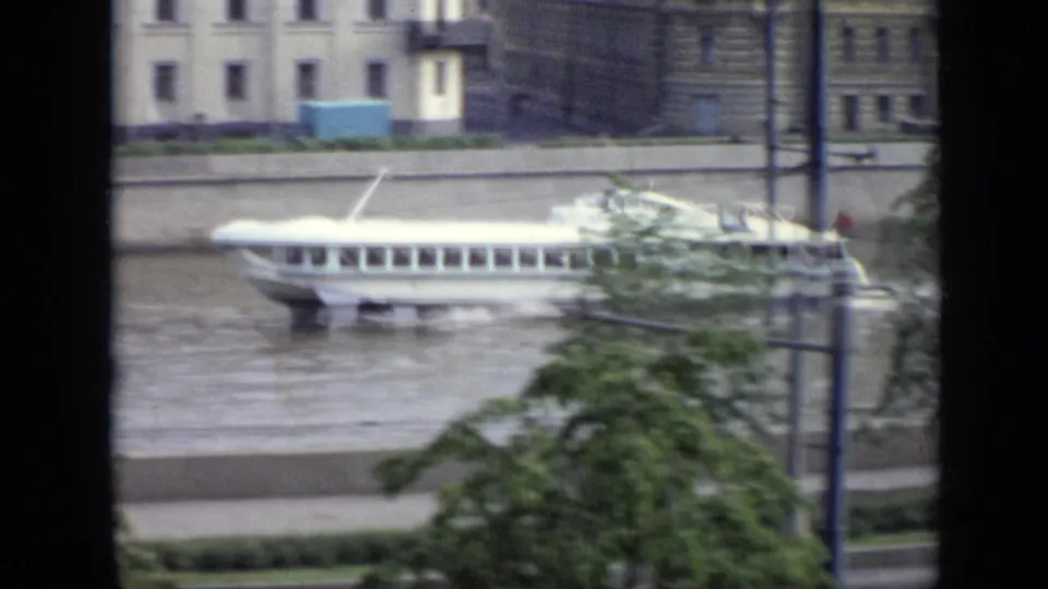 A white passenger riverboat passes along the Moscow embankment, with foliage, poles, and distant pedestrians visible on the riverside path.
