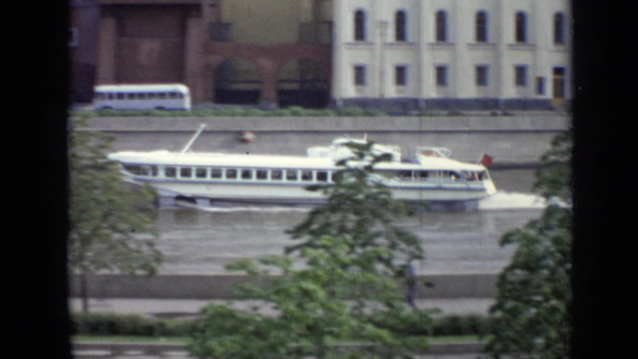 White passenger boat on a river beside embankment buildings and trees, viewed through dark vertical window bars.