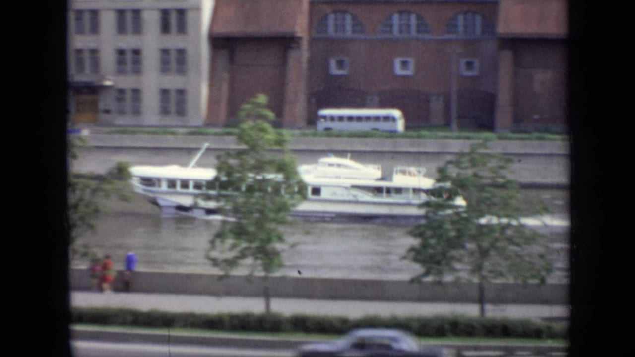 Passenger boat moving along the river with pedestrians, trees, a riverside road, and large buildings behind it.