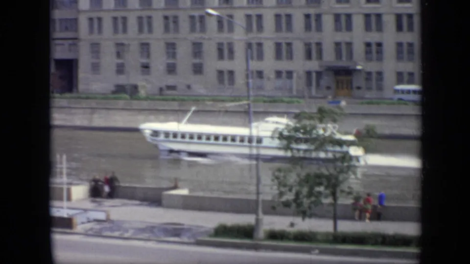 1971 Moscow home-movie frame showing a white river passenger boat, embankment wall, roadside bus, small pedestrians, trees, and urban buildings.