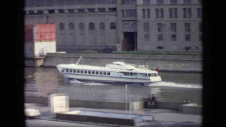 White passenger boat on a Moscow river in 1971, framed by embankment architecture, pedestrians, a small car, trees, and dark foreground bars.