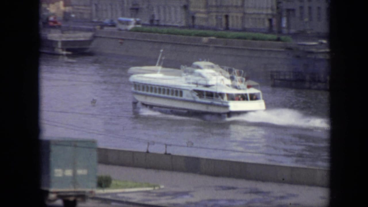 White speedboat crossing a river with buildings and an embankment behind it.