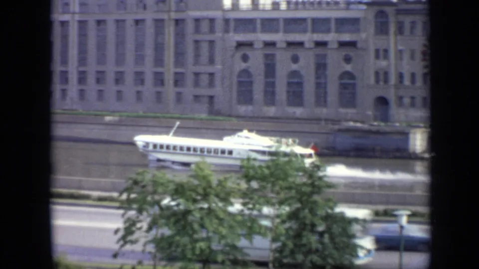 1971 Moscow riverside view with a white passenger boat moving along the water below pale buildings, trees, and roadway traffic space.