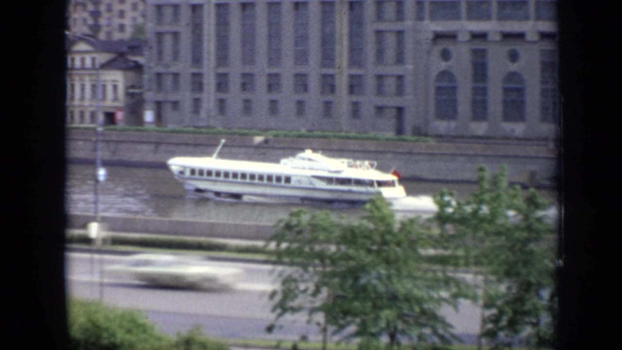 1971 Moscow 8mm frame of a white hydrofoil on the river, seen from above with cars, trees, and riverside buildings nearby.