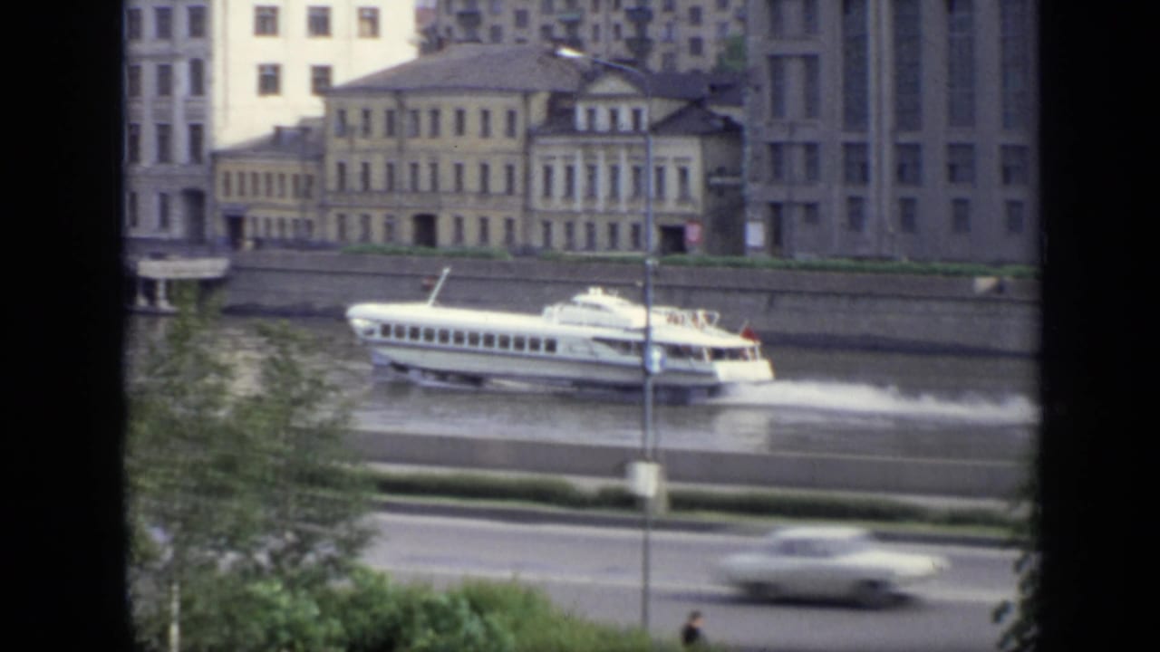 The white hydrofoil continues across the Moscow riverfront, with a stone embankment, traffic, trees, and older buildings in view.