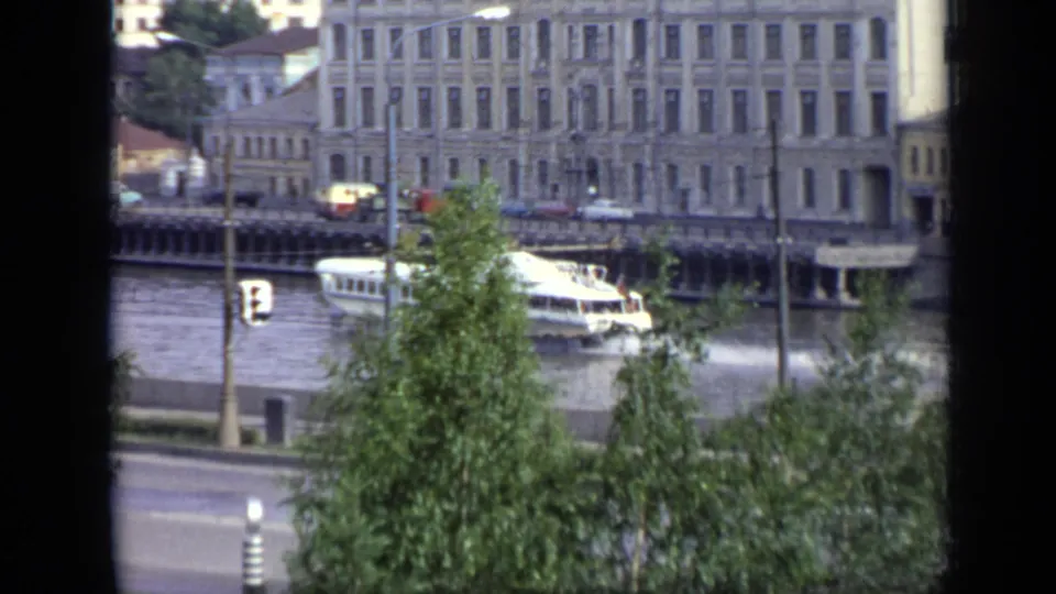 The hydrofoil moves near a riverside structure in Moscow, with leafy trees, traffic lights, road traffic, and building facades visible.