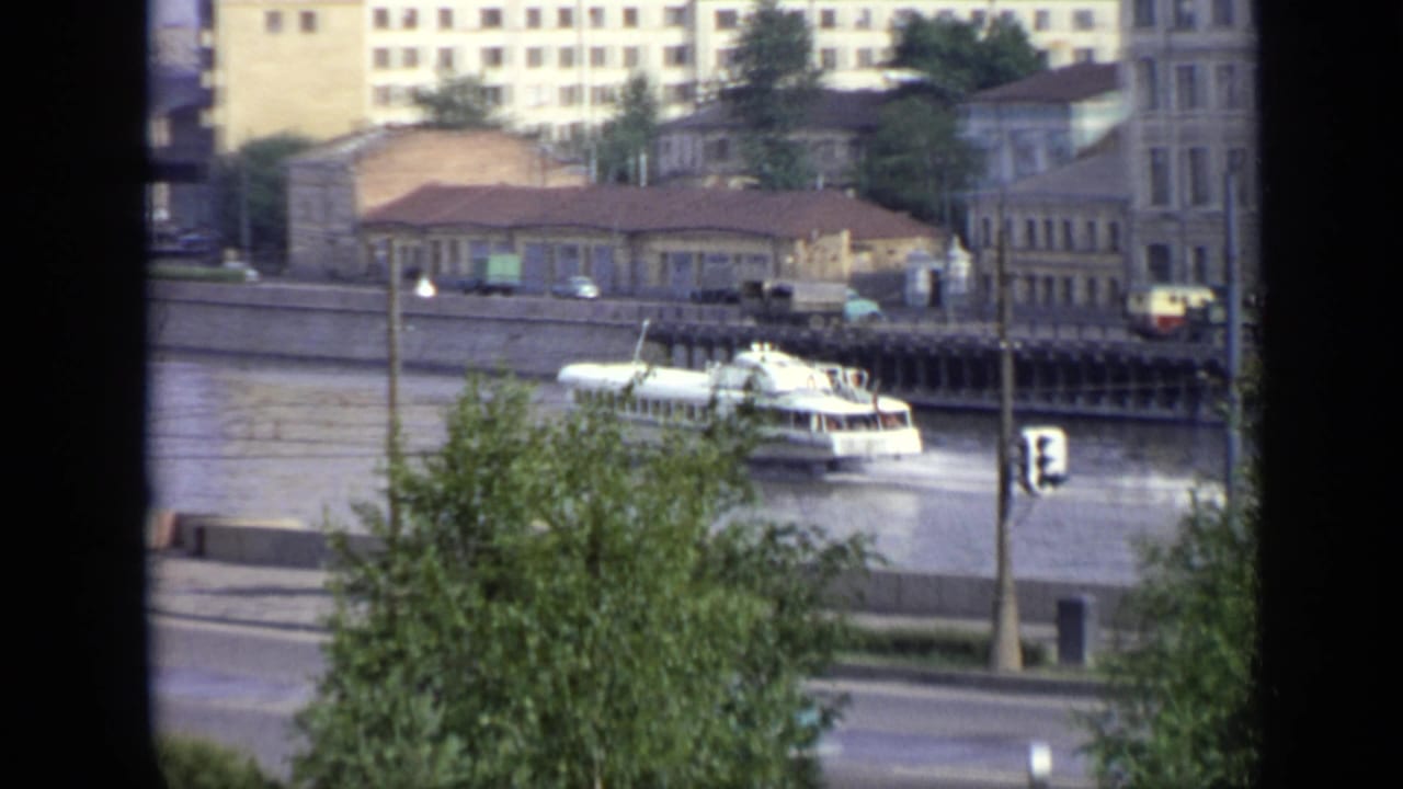 Riverboat near the embankment with trees, traffic signals, and city buildings behind it.