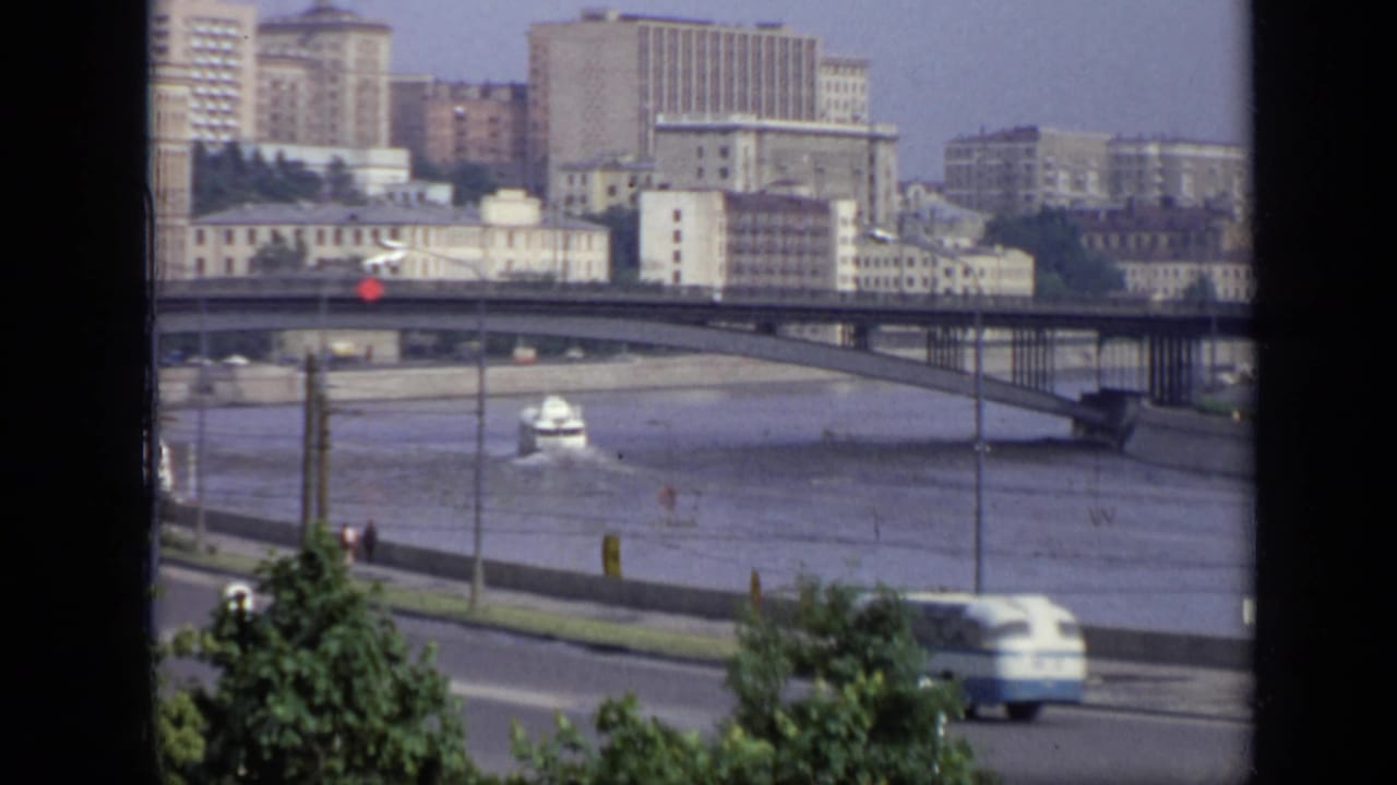 1971 8mm view over a Moscow riverfront, showing a bridge, embankment road, moving traffic, trees, and Soviet-era buildings.