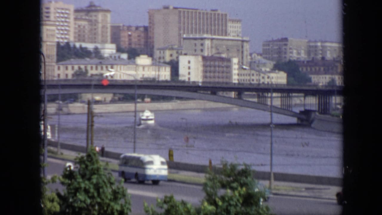 Moscow riverfront in 1971 with a bridge, small boat, roadside bus, trees, and apartment blocks seen from an elevated position.