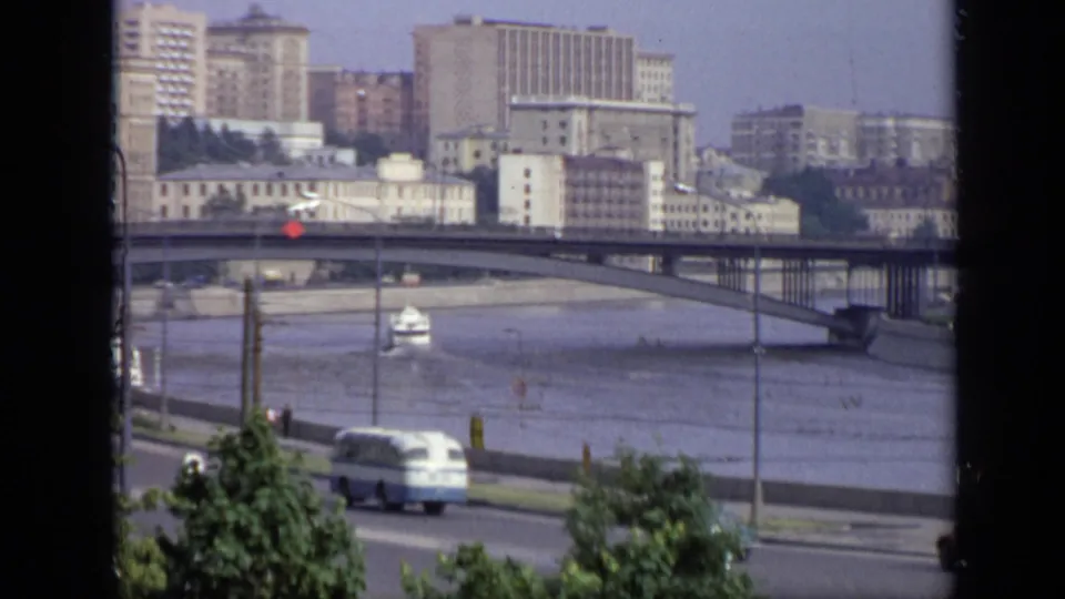 Moscow riverfront in 1971 with a bridge, small boat, roadside bus, trees, and apartment blocks seen from an elevated position.