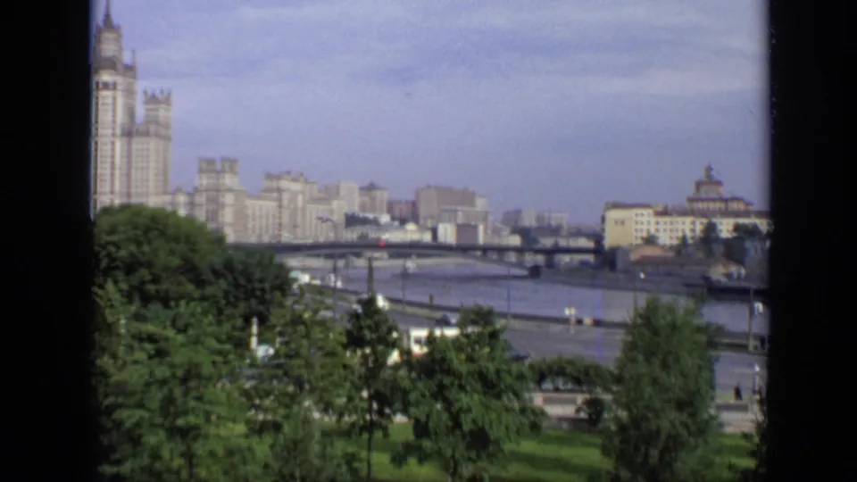 1971 Moscow home-movie frame with trees in the foreground, a river bridge, traffic, apartment blocks, and a tall building on the skyline.