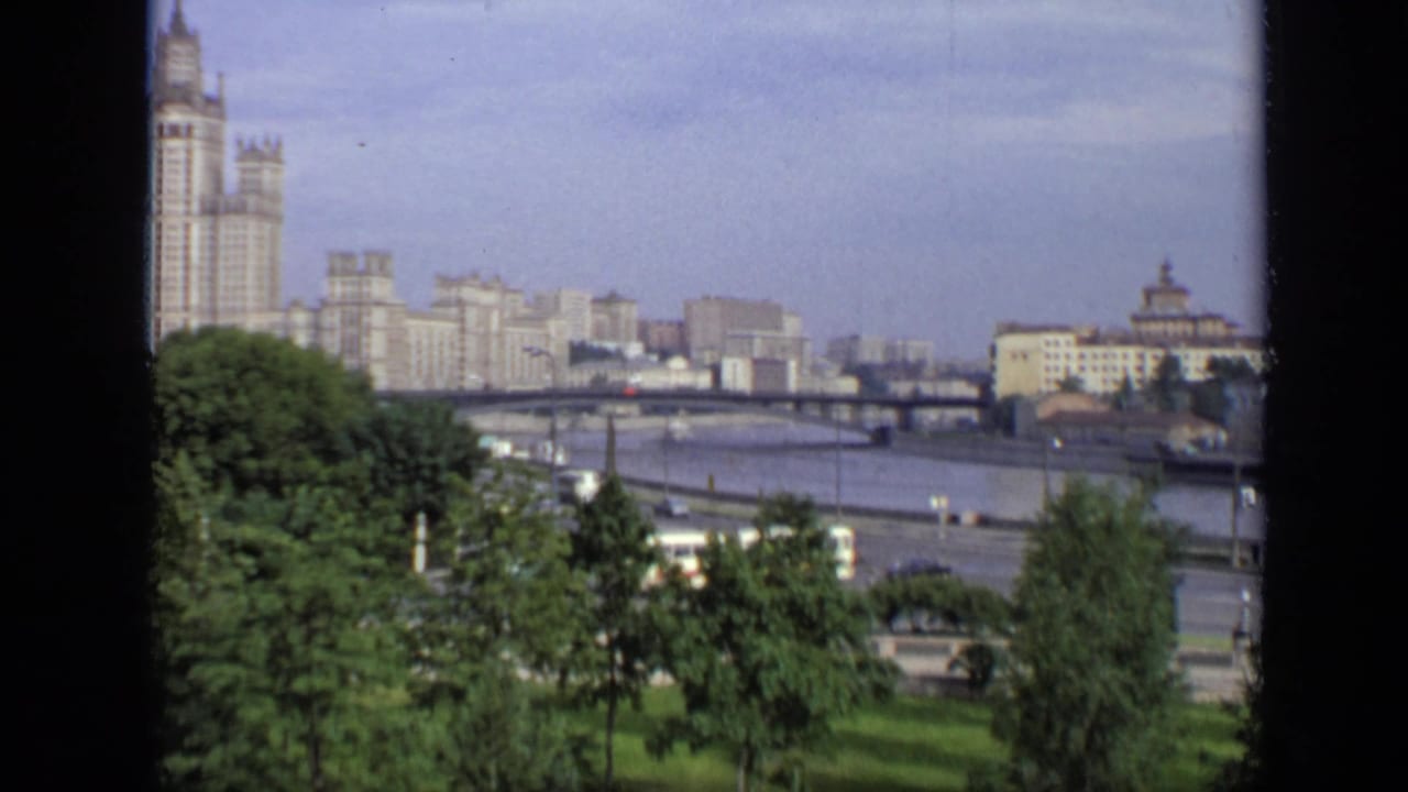 1971 Moscow home-movie frame showing green trees, a river bridge, moving traffic, and a Stalinist high-rise in the distance.