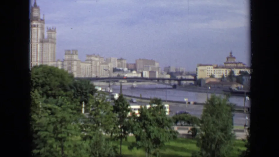 1971 Moscow home-movie frame showing green trees, a river bridge, moving traffic, and a Stalinist high-rise in the distance.