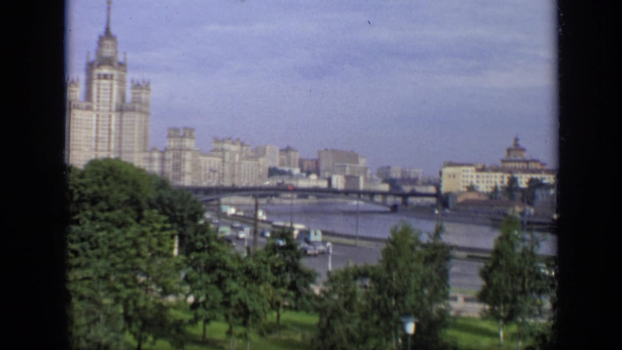Riverfront traffic passes below trees with a bridge and tall pale building beyond.