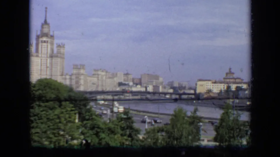 1971 Moscow home-movie view of trees beside the river, a bridge with traffic, and pale high-rise buildings under a clear sky.