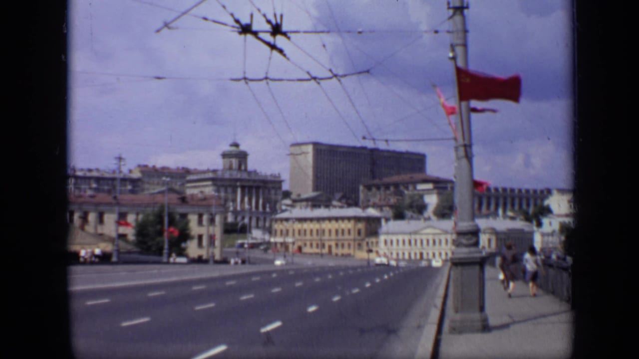 1971 Moscow view across a multi-lane street with red flags, overhead trolley wires, distant vehicles, pedestrians, and large civic buildings.