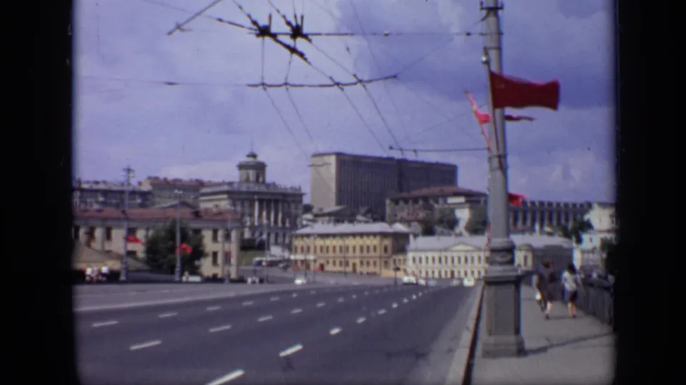 1971 Moscow view across a multi-lane street with red flags, overhead trolley wires, distant vehicles, pedestrians, and large civic buildings.